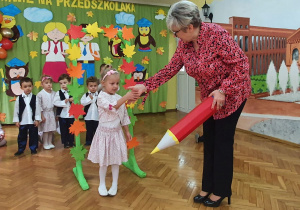 Pani Dyrektor pasuje na przedszkolaka czerwoną kredką dziewczynkę ubraną w białą podkoszulkę oraz spódniczkę i bolerko w kwiatki.
